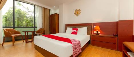 A hotel room with a large bed, wooden furniture, a nightstand with a lamp, and a view of greenery through the window.