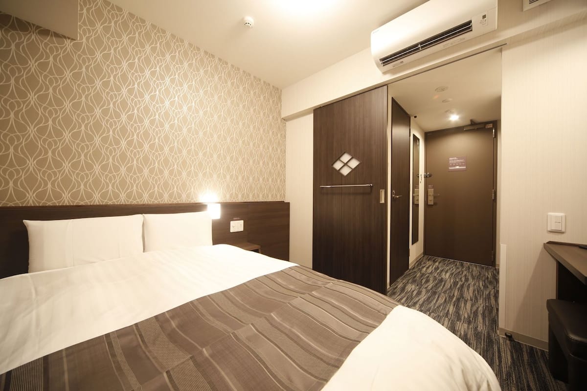 A hotel room with a bed, a wardrobe, an air conditioning unit, and a door leading to a bathroom.