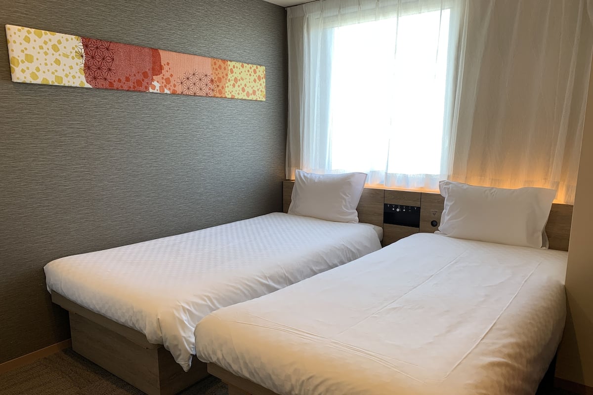 Ein Hotelzimmer mit zwei Einzelbetten, einem großen Fenster mit transparenten Vorhängen und einem strukturierten Wandbild über den Betten.