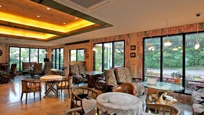 A spacious indoor area with multiple seating arrangements, large windows offering views of greenery, and a mix of wooden and upholstered furniture. - The Areumdaun Pension (Seogwipo)