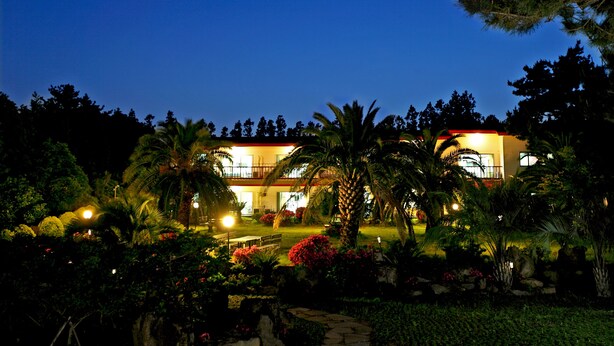 A well-lit building with a balcony, surrounded by lush greenery and flowering plants. - The Areumdaun Pension (Seogwipo)