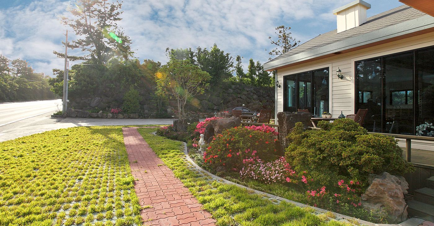 A residential property with a paved driveway, a garden with various plants and flowers, a patio area with outdoor furniture, and a large windowed building.