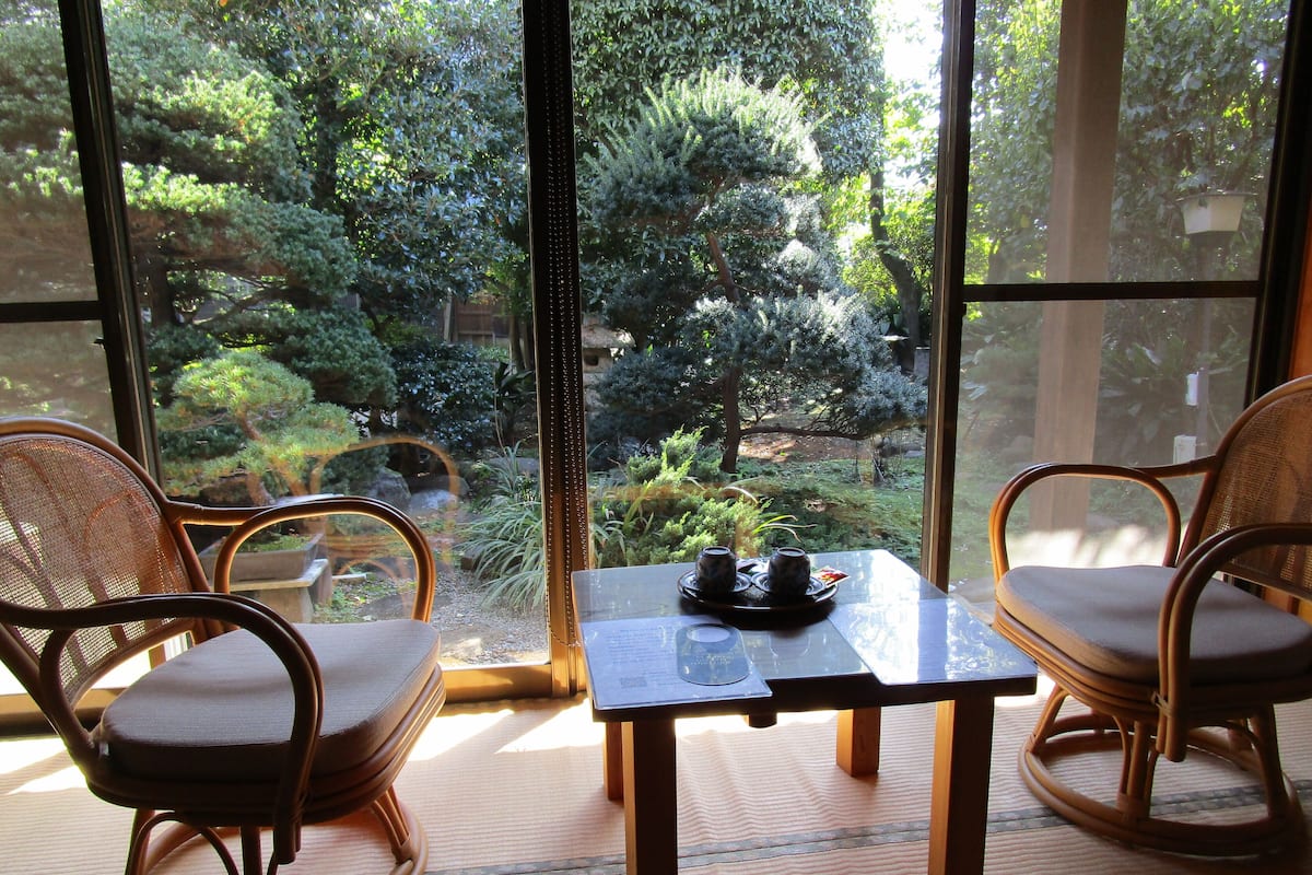Quarto com duas cadeiras de vime, uma mesa pequena e vista para um jardim através de janelas amplas.