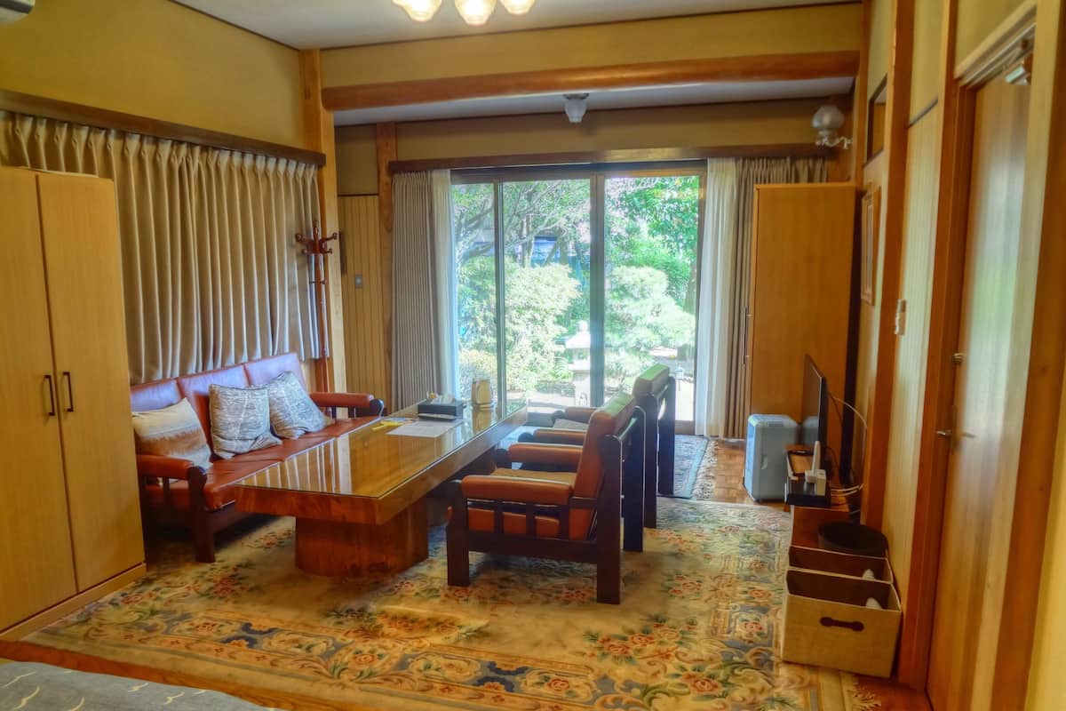 Uma sala de estar acolhedora com sofá, cadeiras, mesa de centro e televisão. Há uma janela grande com cortinas, uma porta e um tapete estampado.