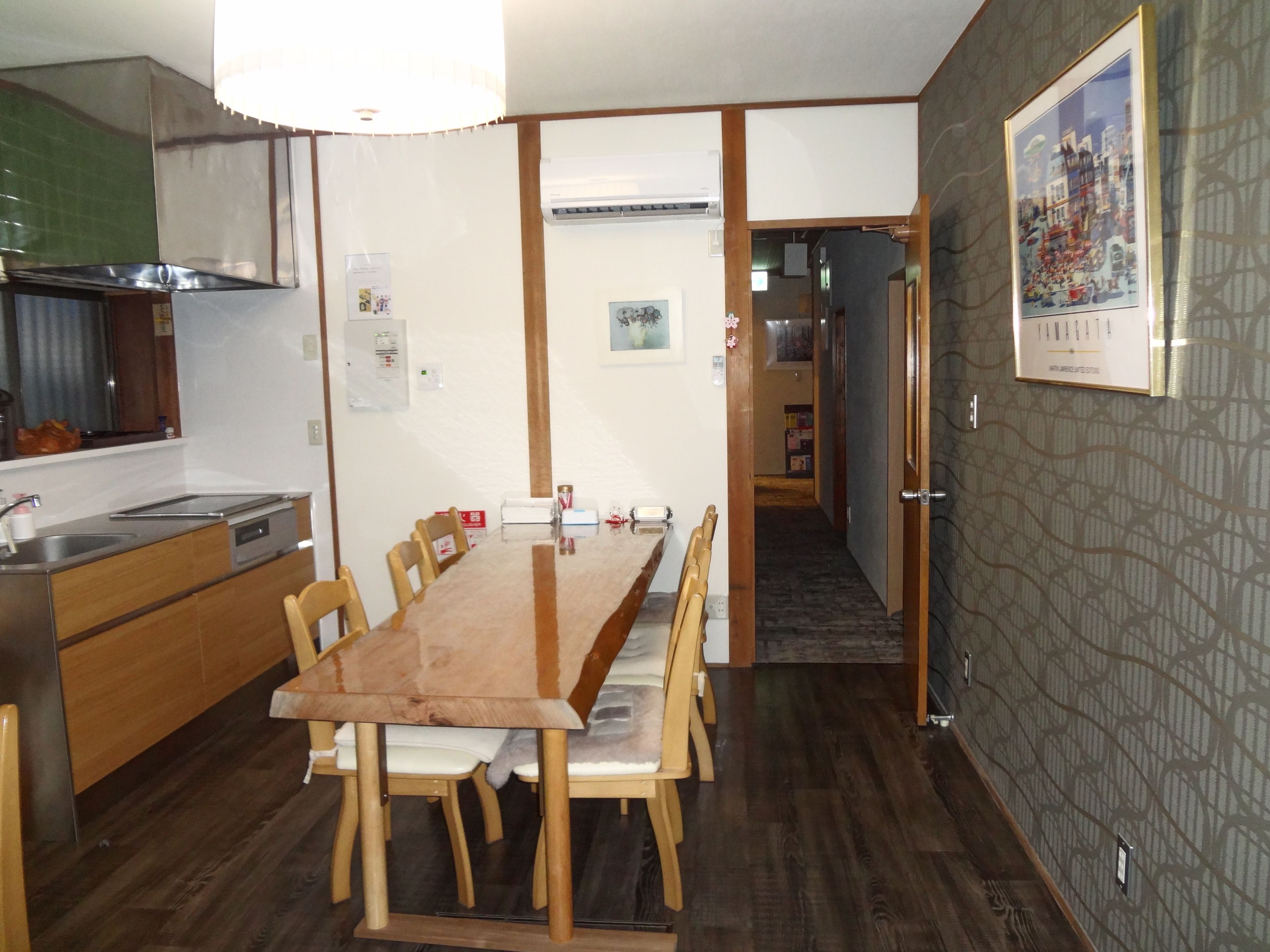 Un comedor con una larga mesa de madera y sillas, una cocina con estufa y lavamanos, y un pasillo que conduce a otra habitación.
