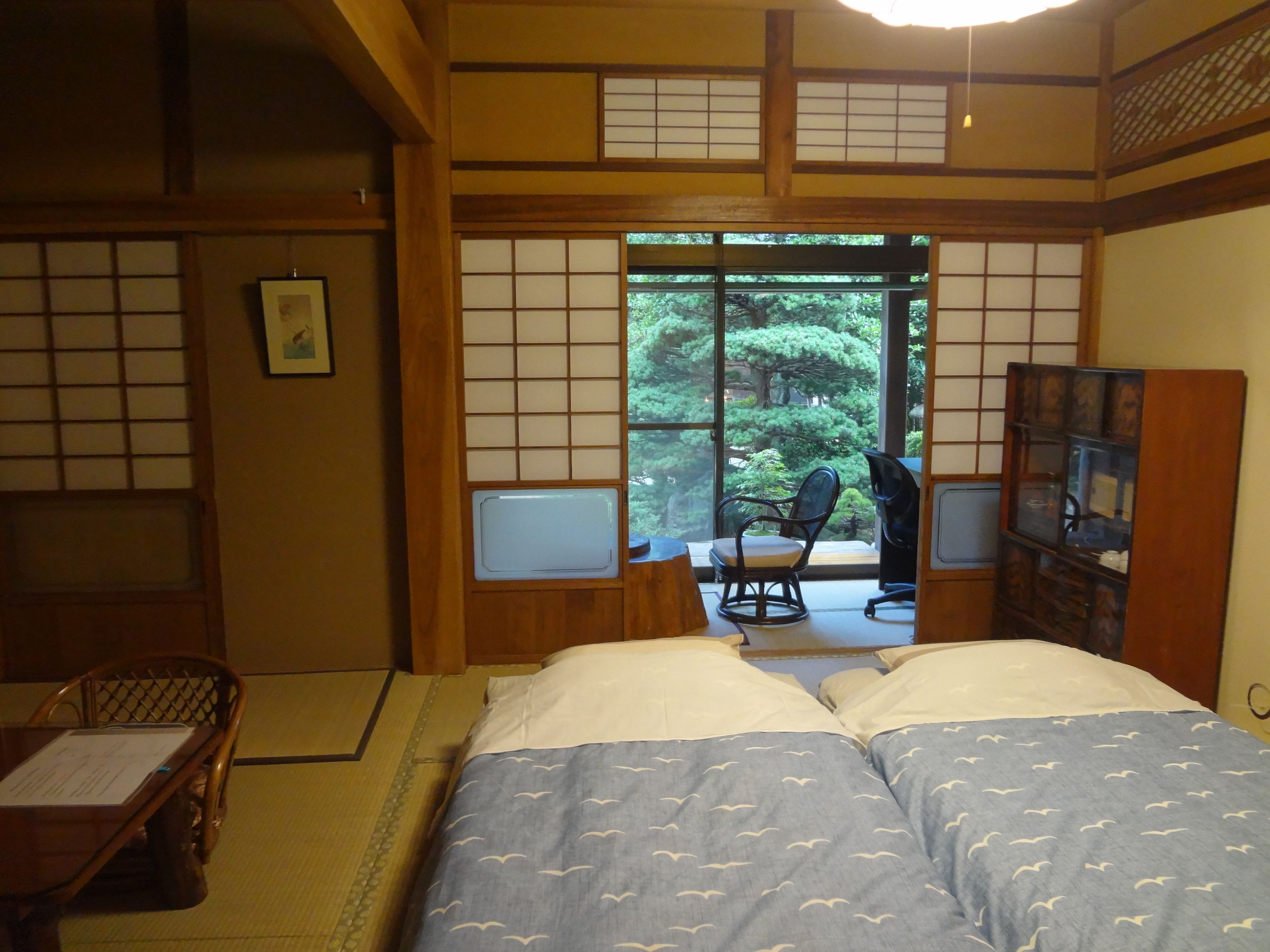 Een traditionele Japanse kamer met tatami vloeren, een bed met een blauwe sprei, een houten bureau en uitzicht op een weelderige tuin door een schuifdeur.