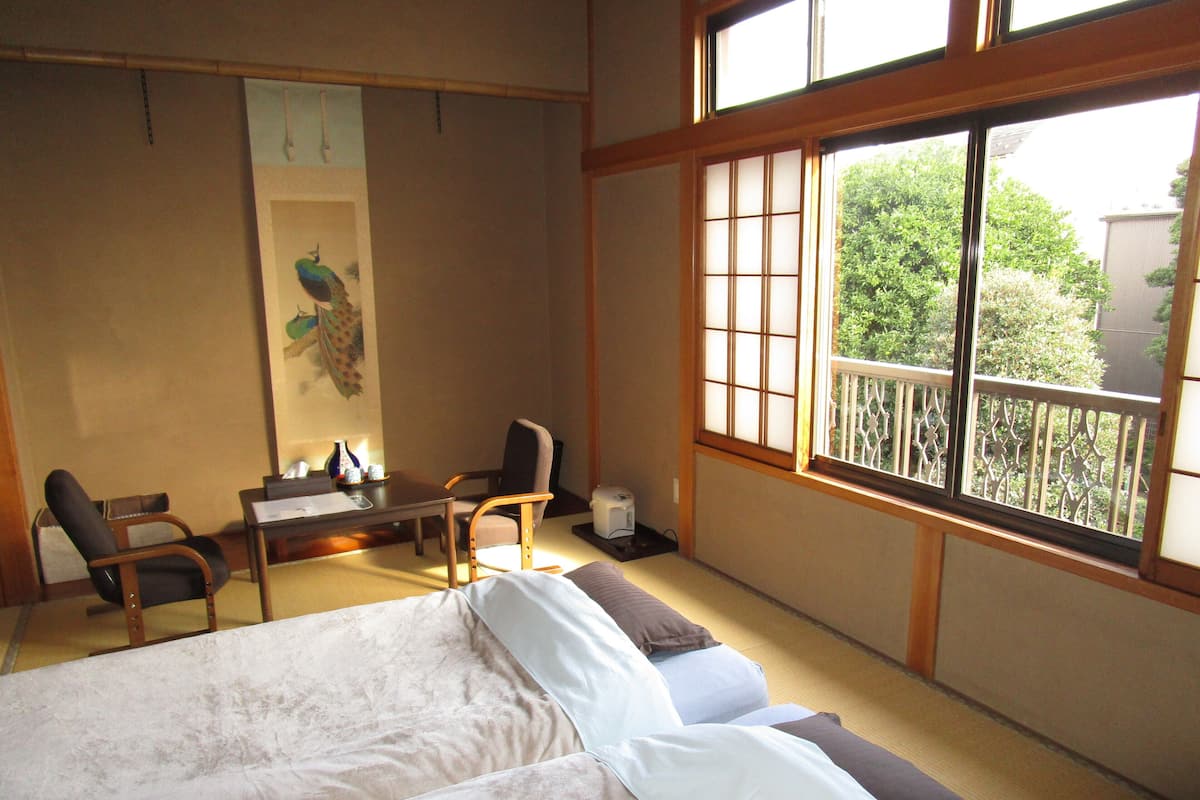 Um quarto japonês tradicional com tatames, uma mesa baixa com cadeiras, uma cama com almofadas e uma janela grande com vista para vegetação.