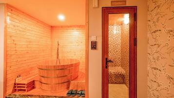A sauna room with a wooden tub, a control panel, and slippers on the floor.