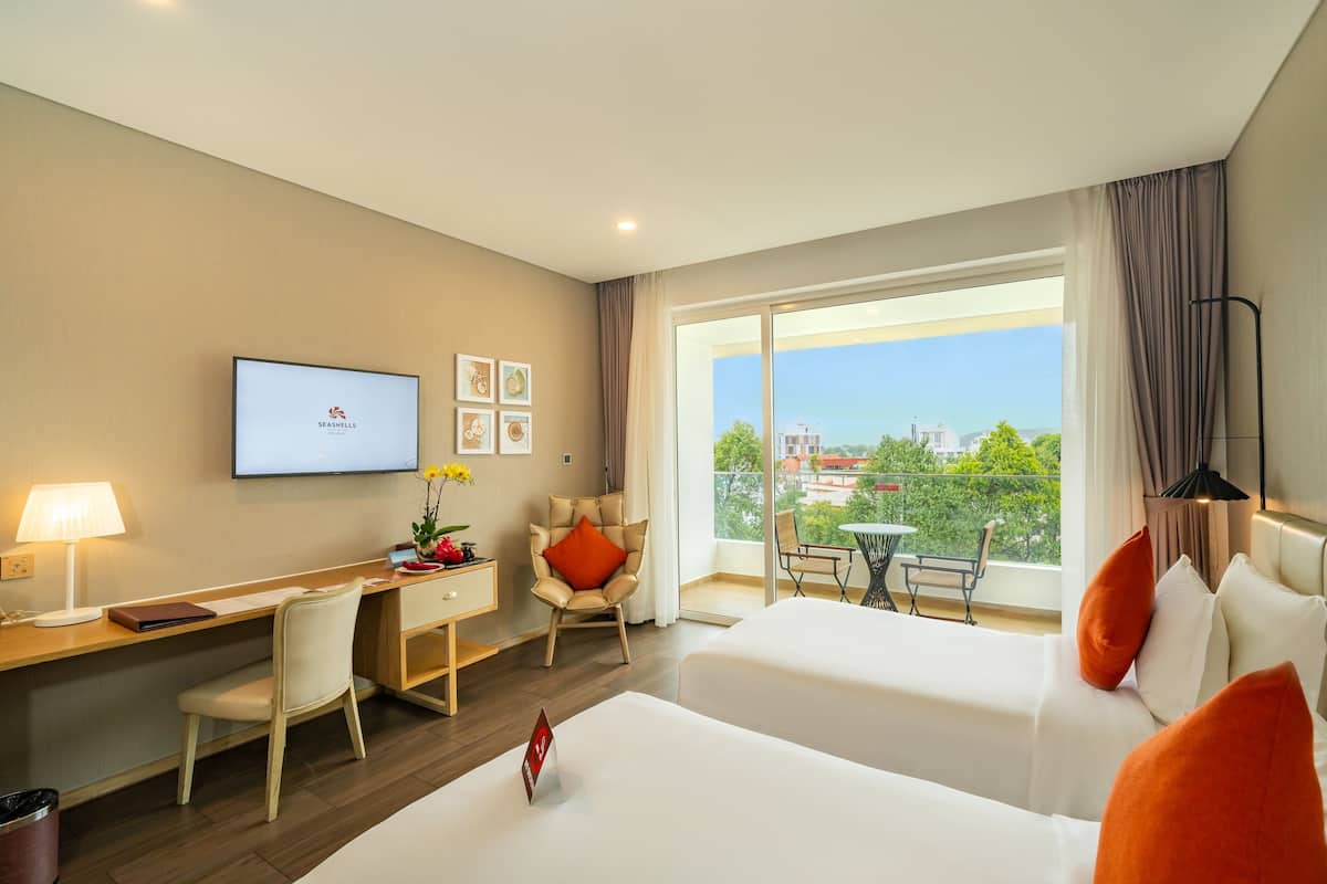 A hotel room with a desk, two beds, a TV, and a balcony with a city view.