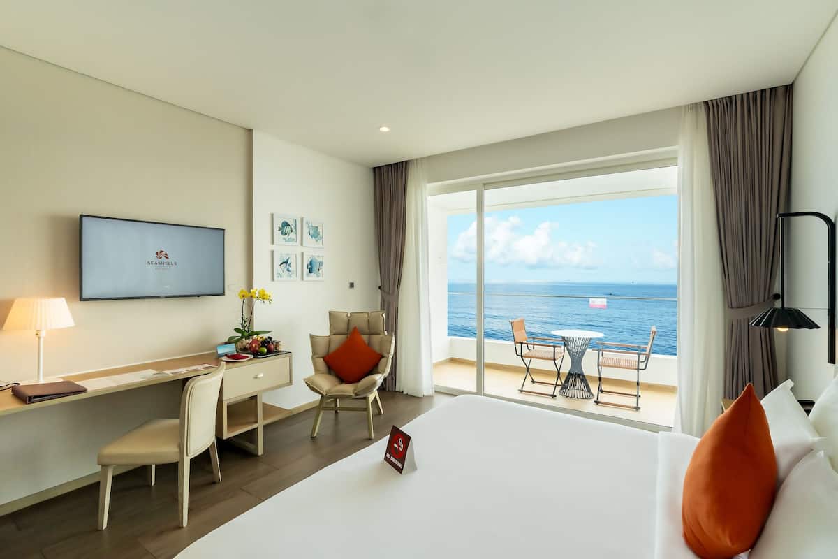 A hotel room with a desk, chair, bed, and a large window offering a view of the sea.