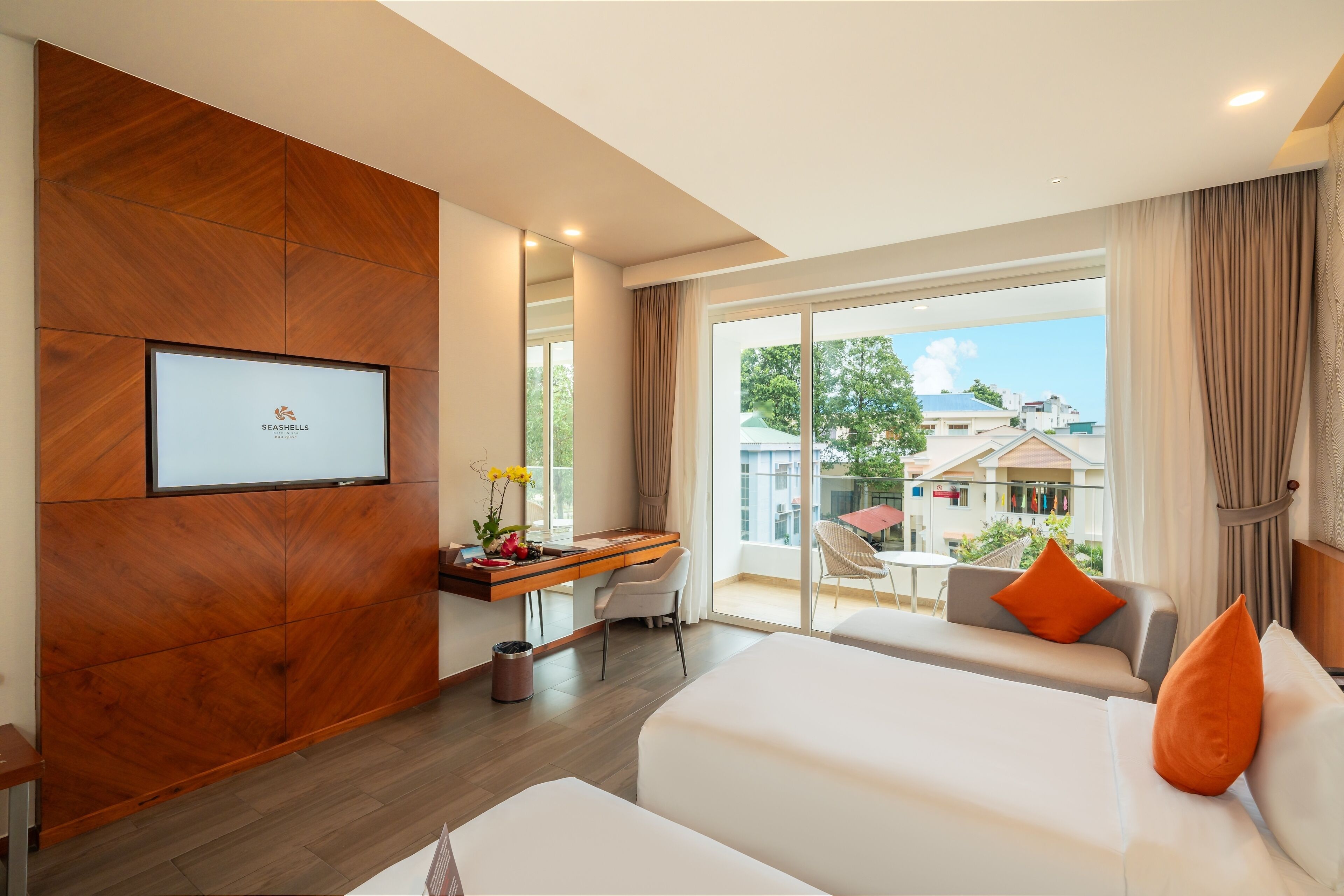 A modern hotel room with a wooden accent wall, a desk with a chair, a bed with orange pillows, and a large window offering a view of the city.