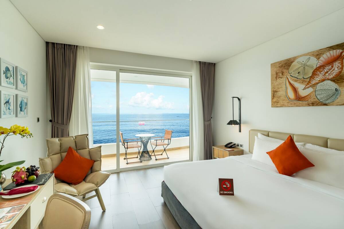A hotel room with a large bed, a desk with a chair, a dining table with chairs, and a balcony with a view of the sea.