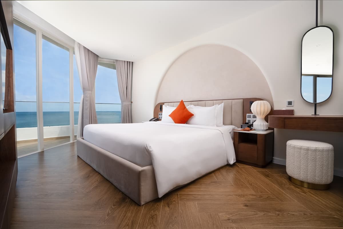 A modern hotel room with a large bed, a nightstand, a round mirror, and a view of the sea.