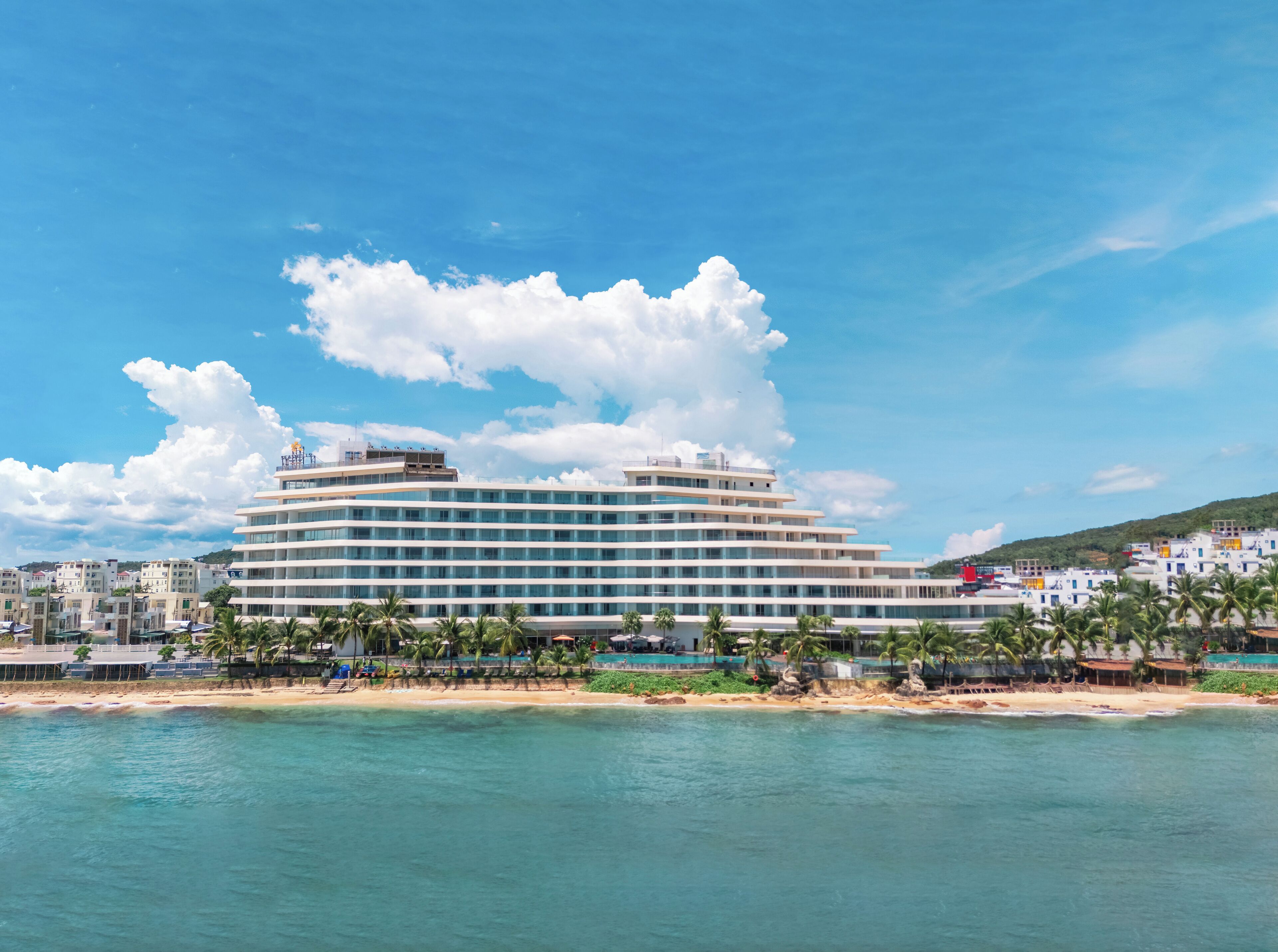 A large, multi-story hotel with a modern design, located near a beach with clear blue waters.