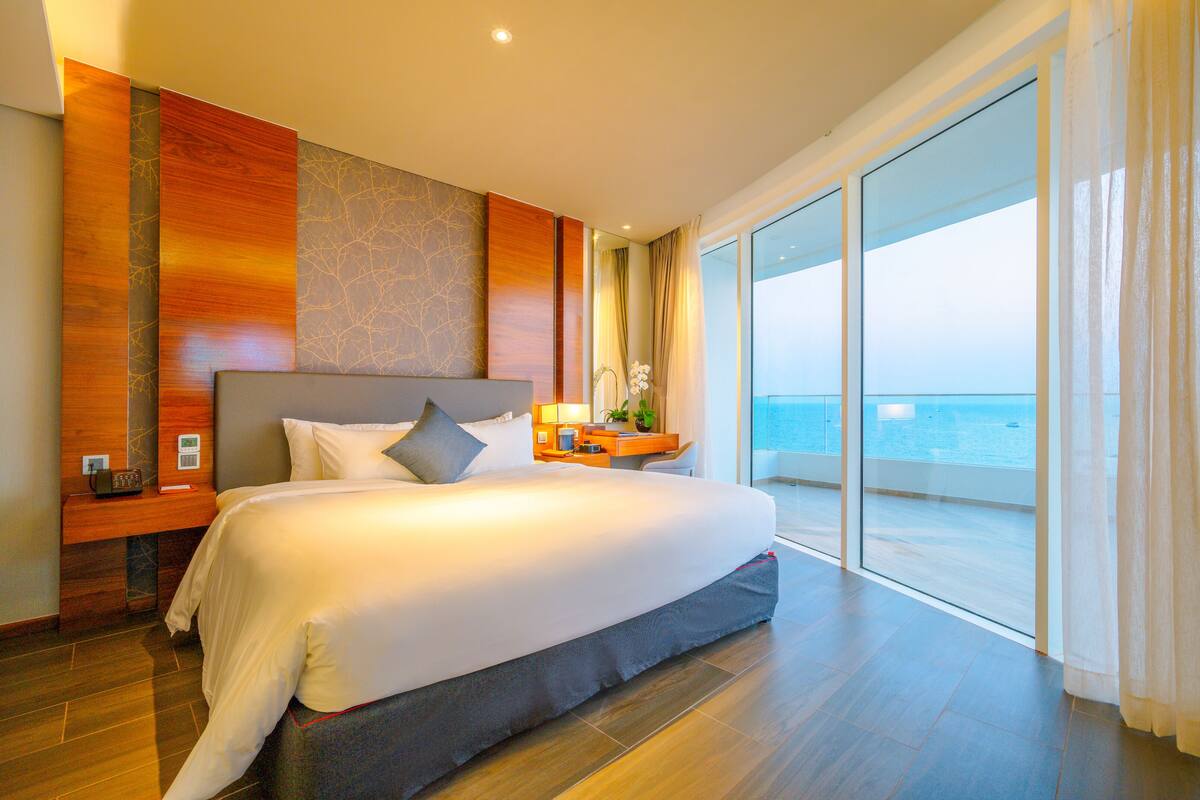 A spacious bedroom with a large bed, a nightstand, and a view of the sea through floor-to-ceiling windows.