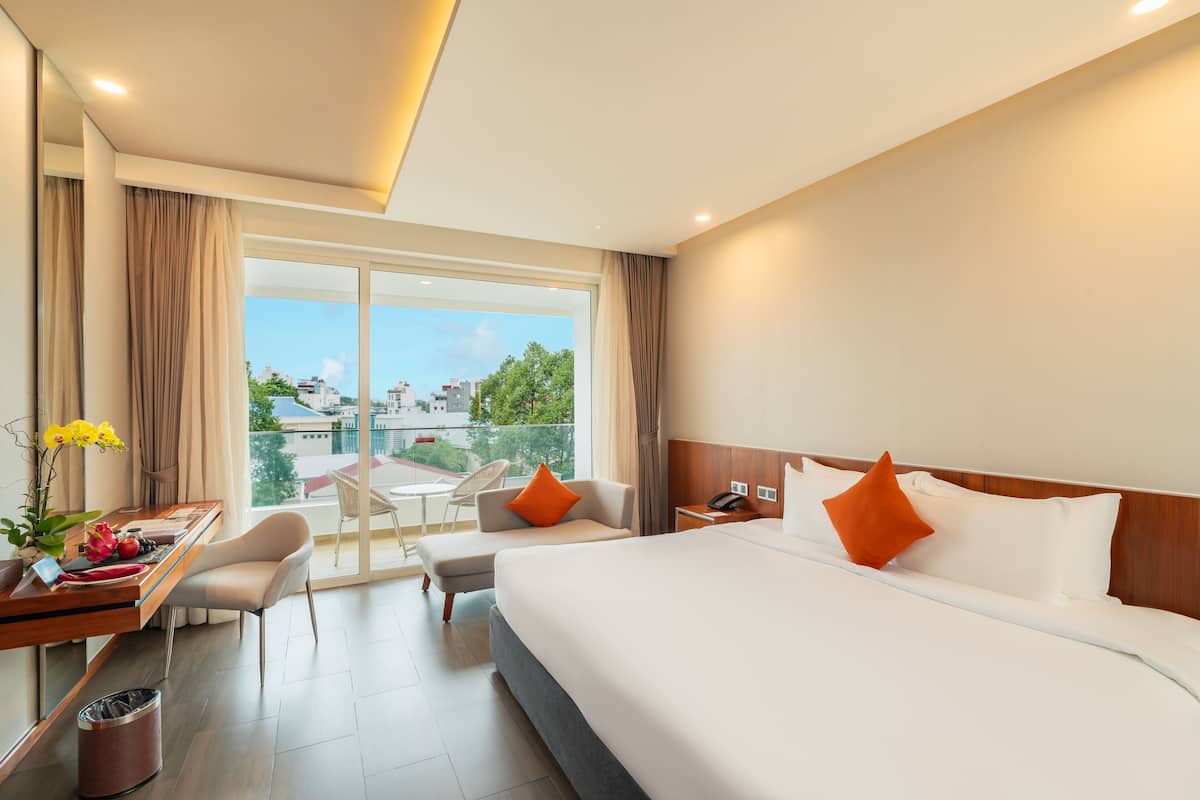 A hotel room with a large bed, two chairs, a desk with a trash bin, and a view of the city through large windows.