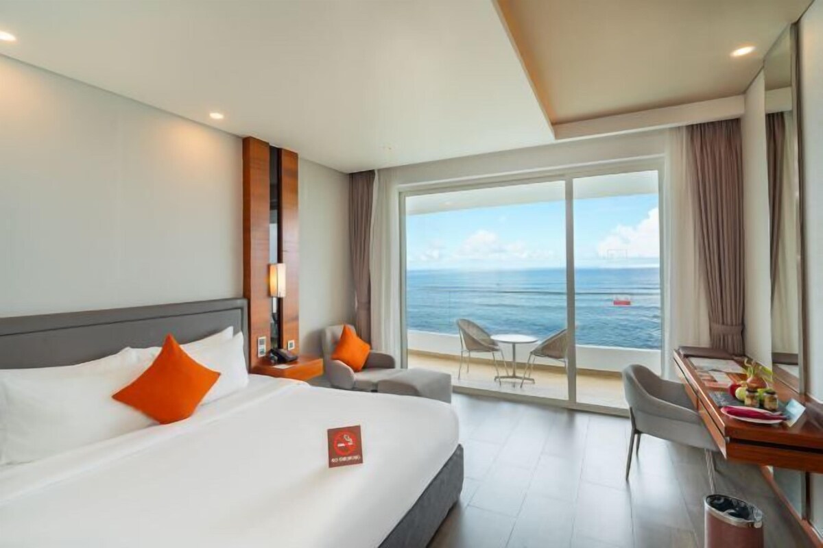 A hotel room with a bed, a desk, a chair, and a view of the sea.
