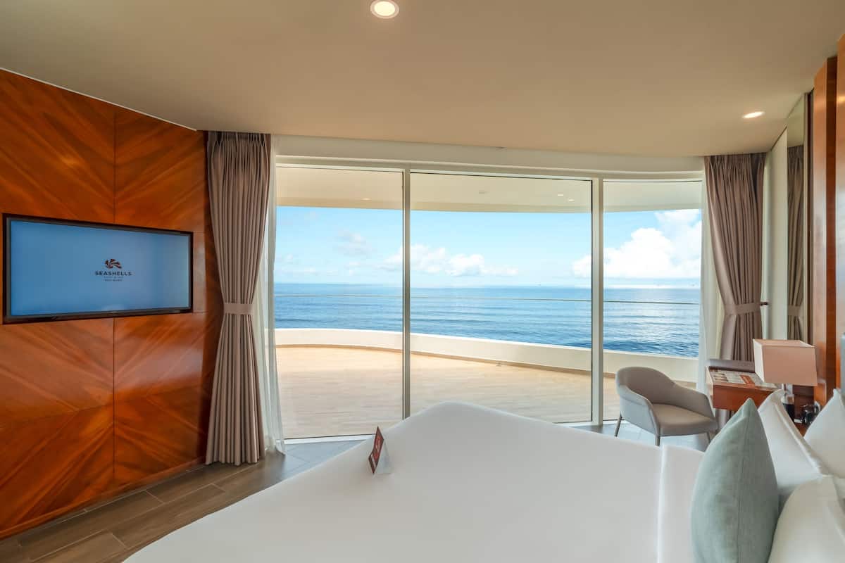 A hotel room with a large bed, a television, a desk, and a view of the ocean.