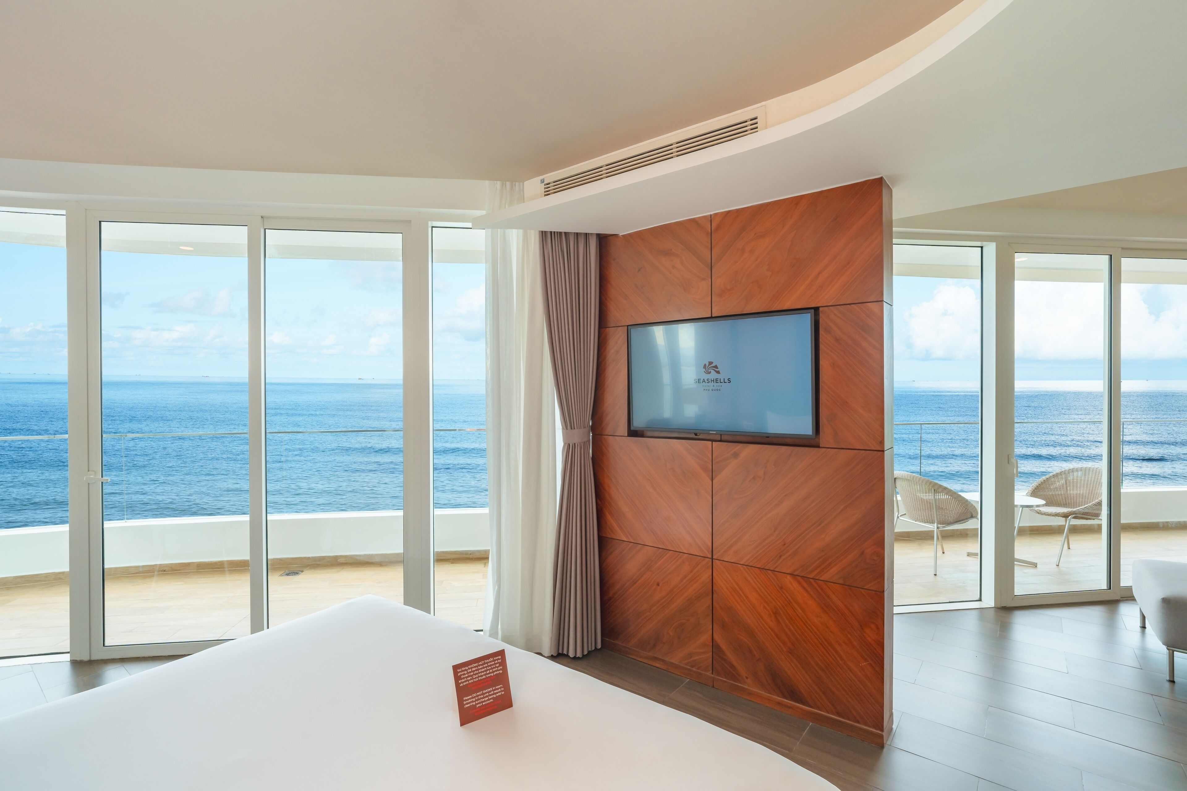 Quarto de hotel moderno com uma cama grande, televisão de tela plana fixada em um painel de madeira e uma varanda com vista para o mar.