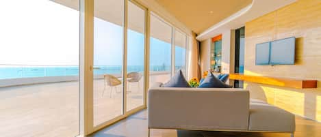 A modern living room with a large sofa, a flat-screen TV mounted on the wall, and a view of the ocean through floor-to-ceiling windows.