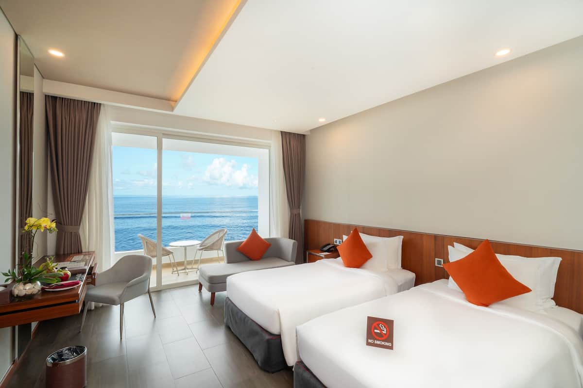 A hotel room with two beds, a desk with a chair, a small table with a vase, and a view of the ocean.