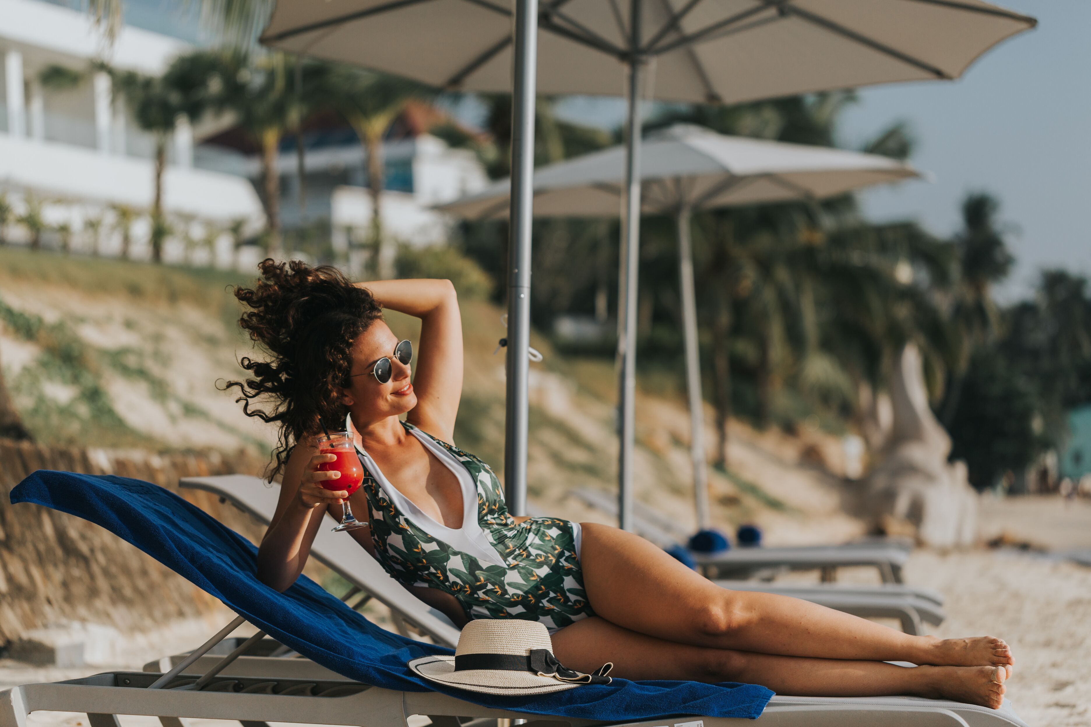 A person relaxing on a beach chair with a drink and a hat, under a beach umbrella.