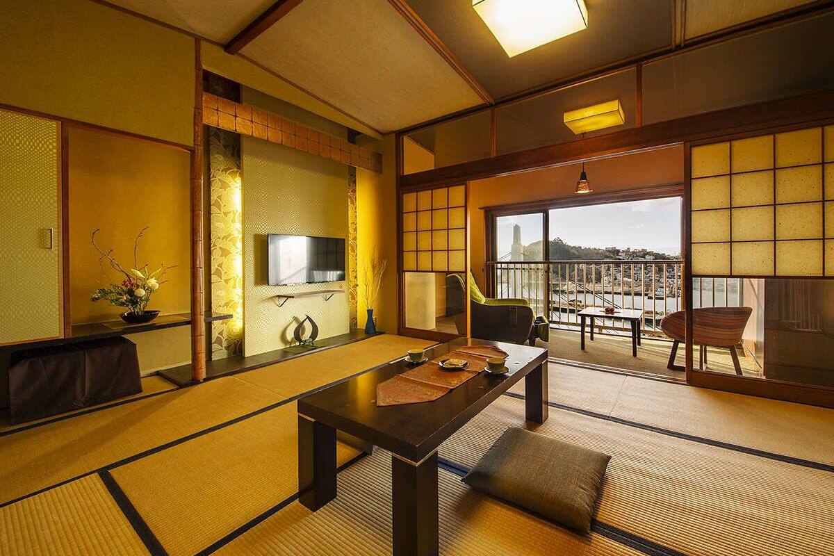 Una habitación tradicional japonesa con suelo de tatami, una mesa baja con cojines, un televisor y vistas a un paisaje urbano a través de amplias ventanas.
