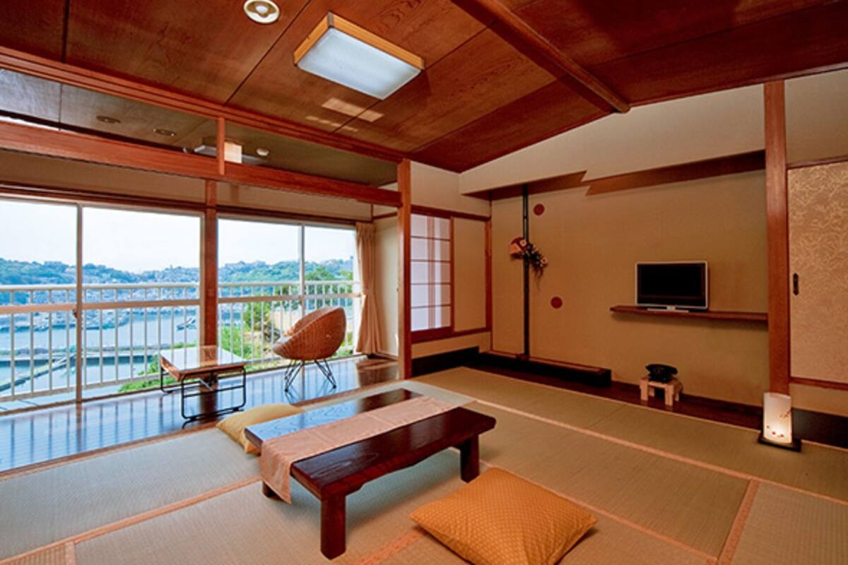 Una habitación japonesa tradicional con suelo de tatami, una mesa de madera baja, una silla acolchada y un televisor en la pared.