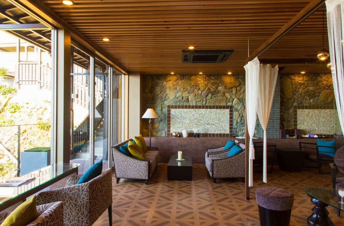 A modern lounge area with patterned sofas, a stone accent wall, and a glass railing overlooking a garden.