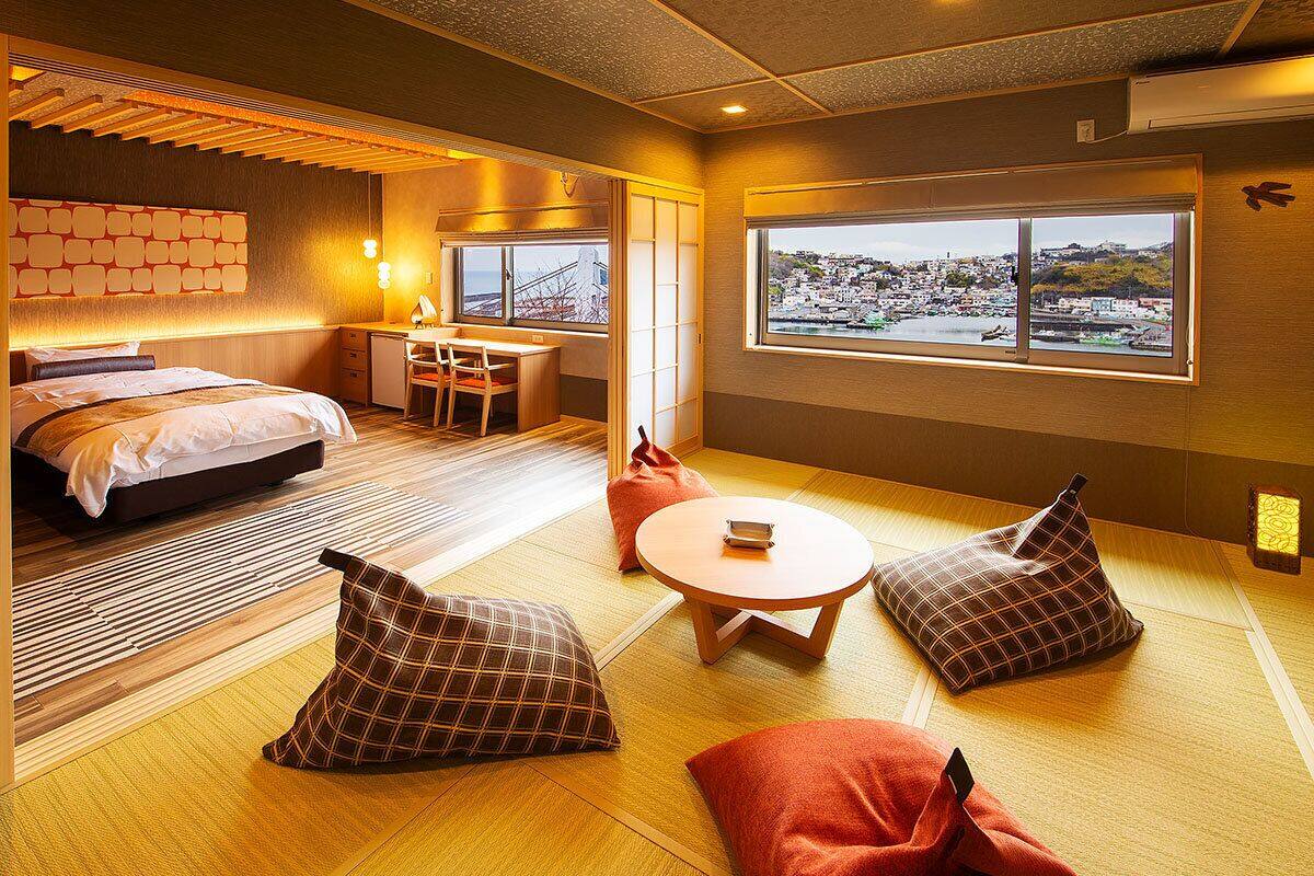 Habitación de hotel con cama, escritorio, silla, mesa redonda y vistas a la ciudad a través de amplias ventanas.