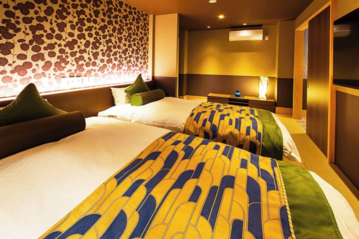 Habitación de hotel con dos camas, un papel tapiz estampado y una mesita de noche con lámpara.