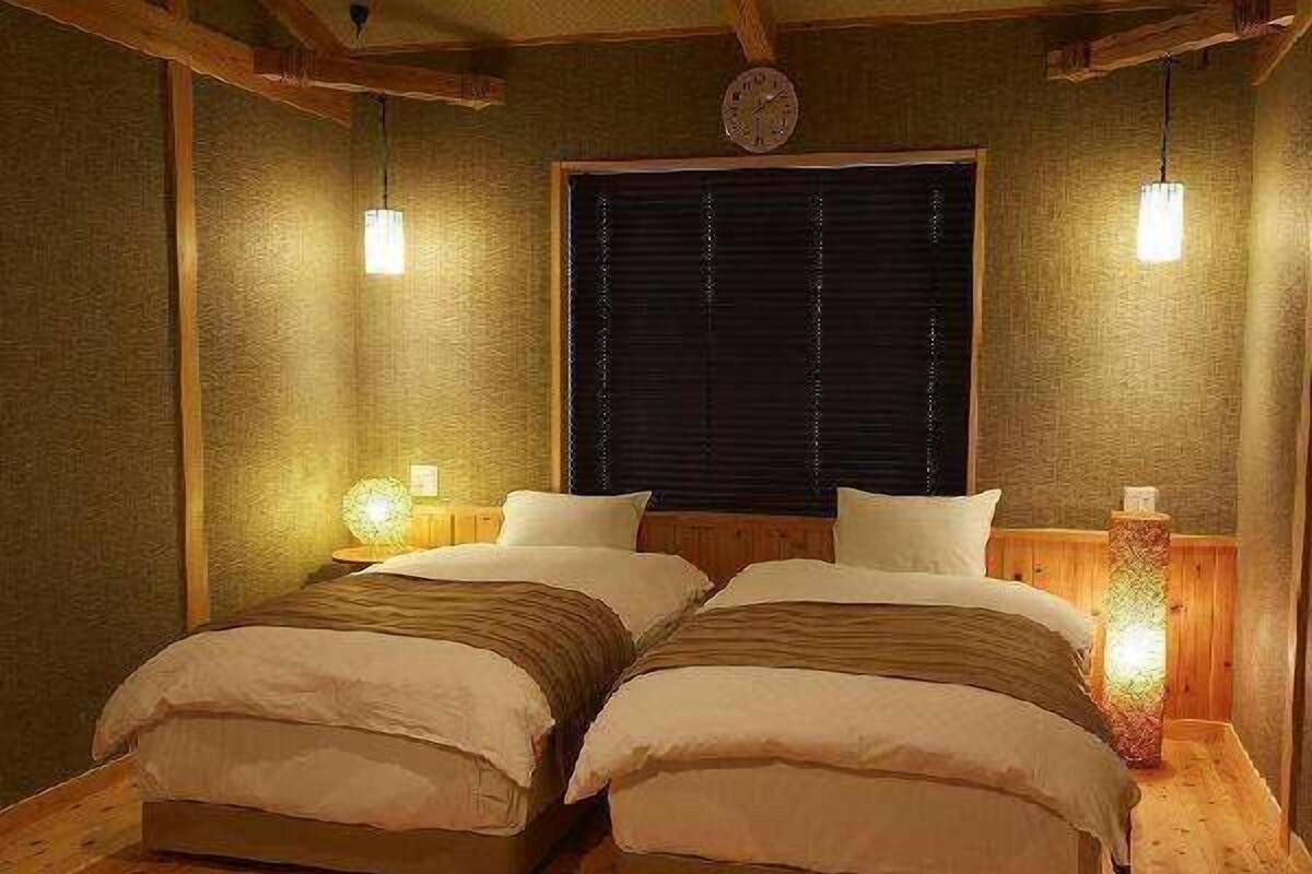 Una habitación con dos camas, vigas de madera, un ventanal con persianas y lámparas fijadas a la pared.