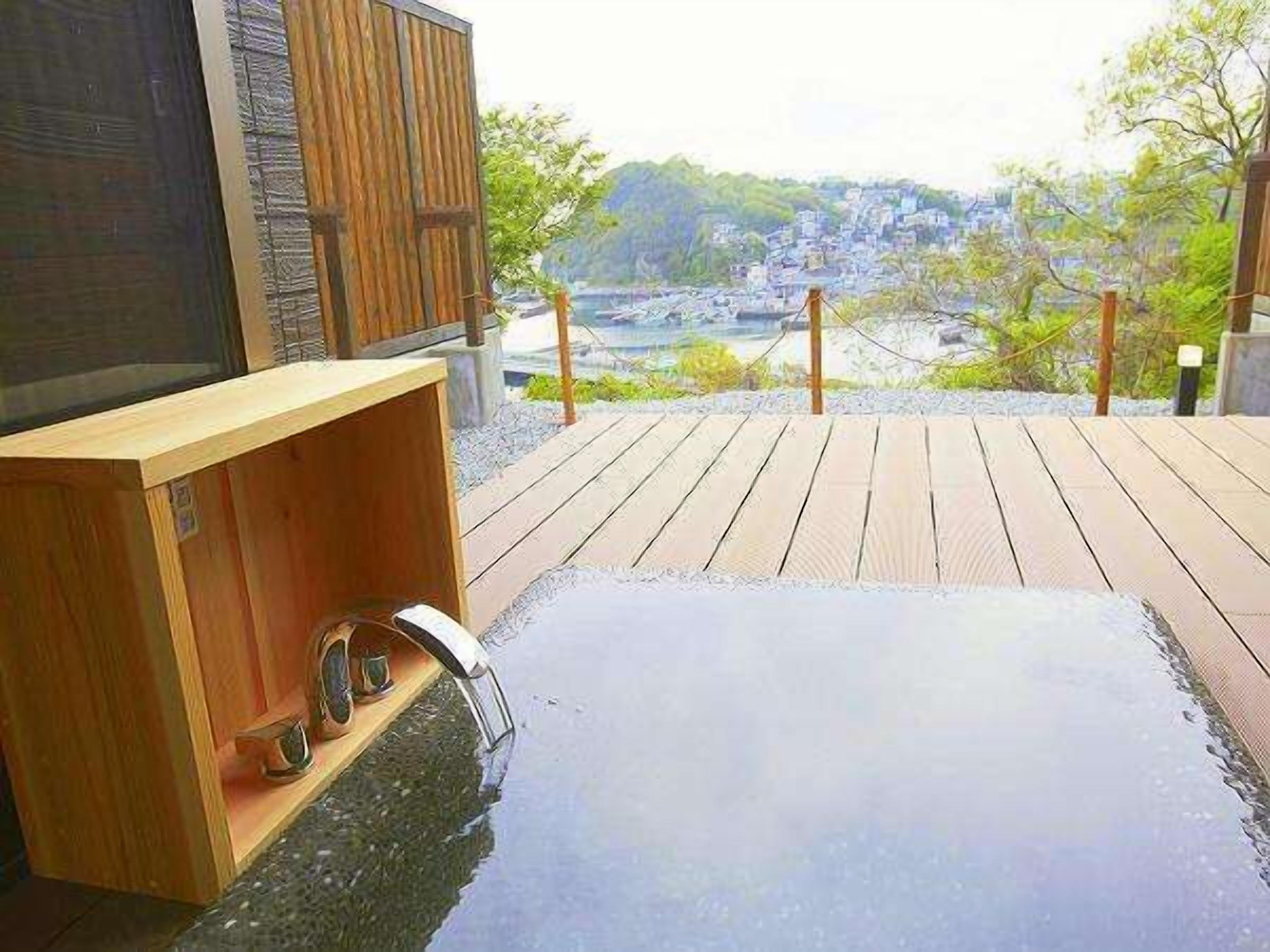 Une terrasse en bois dotée d’un jacuzzi et d’une structure en bois, offrant une vue panoramique sur un paysage pittoresque, avec des bâtiments et de la végétation.