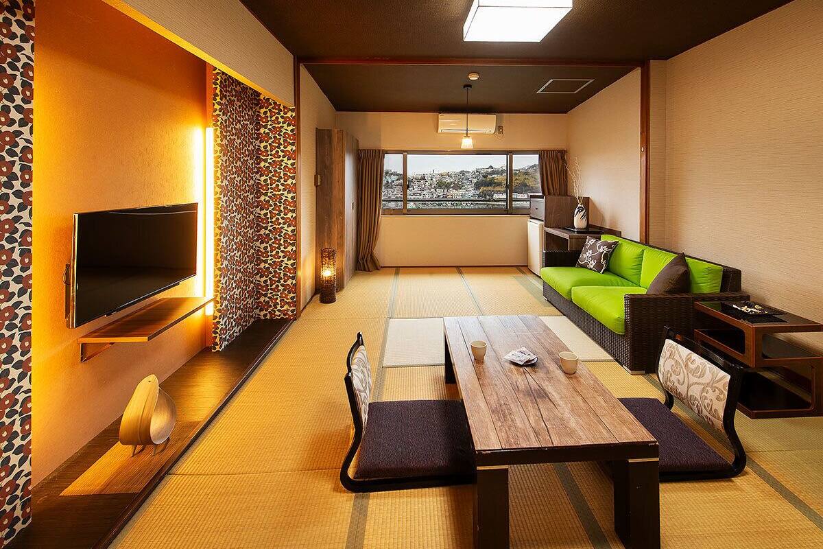 Una habitación tradicional japonesa con suelo de tatami, una mesa baja con cojines, un sofá verde y un televisor en la pared izquierda.