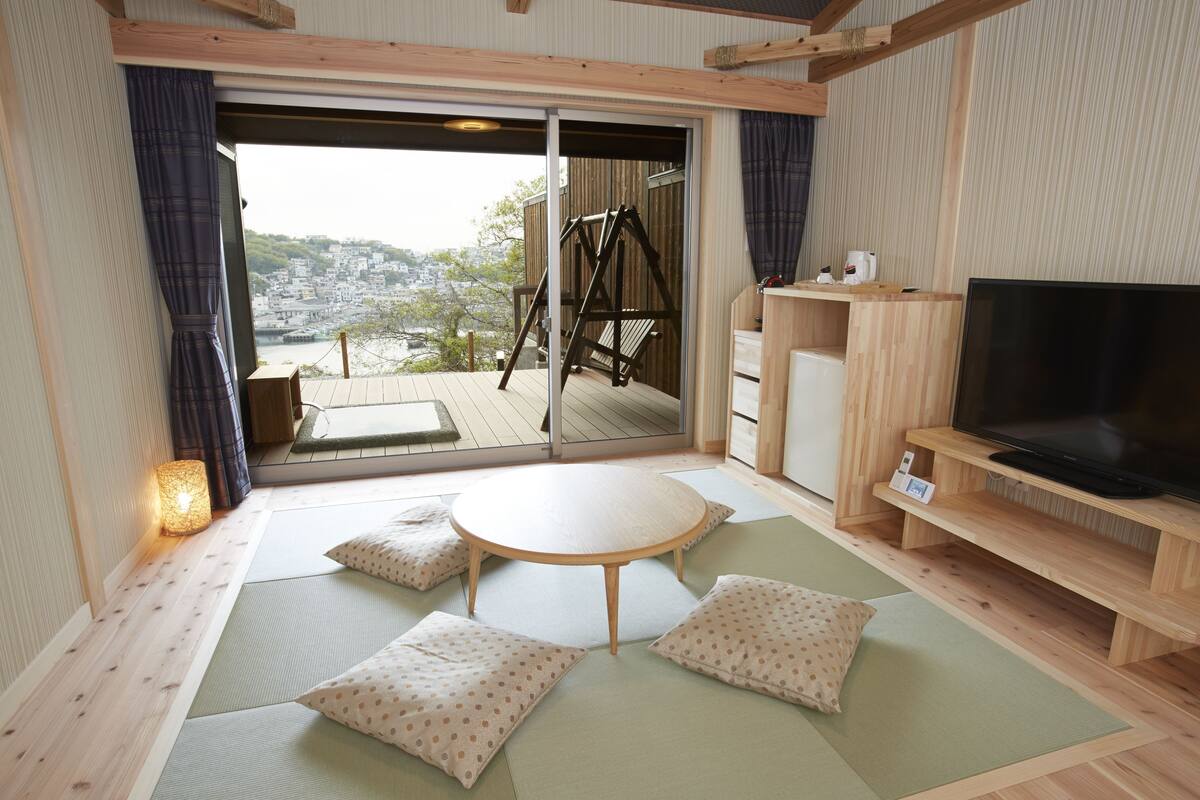 Una habitación japonesa tradicional con suelo de tatami, una mesa de madera baja, cojines, un televisor y una puerta corrediza de cristal que da a una terraza exterior.