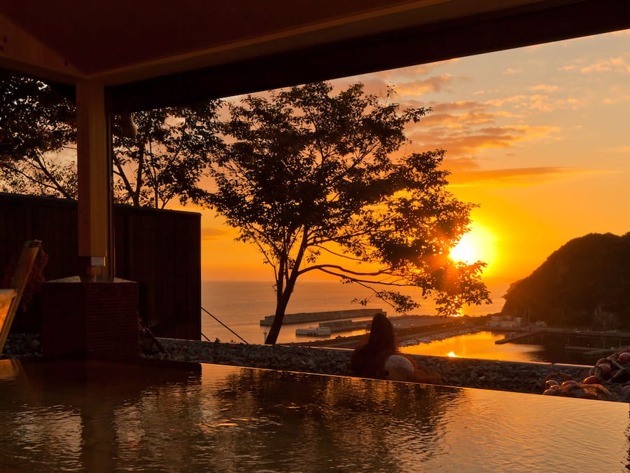 A room with a view of a sunset over water, a tree, and a stone wall.
