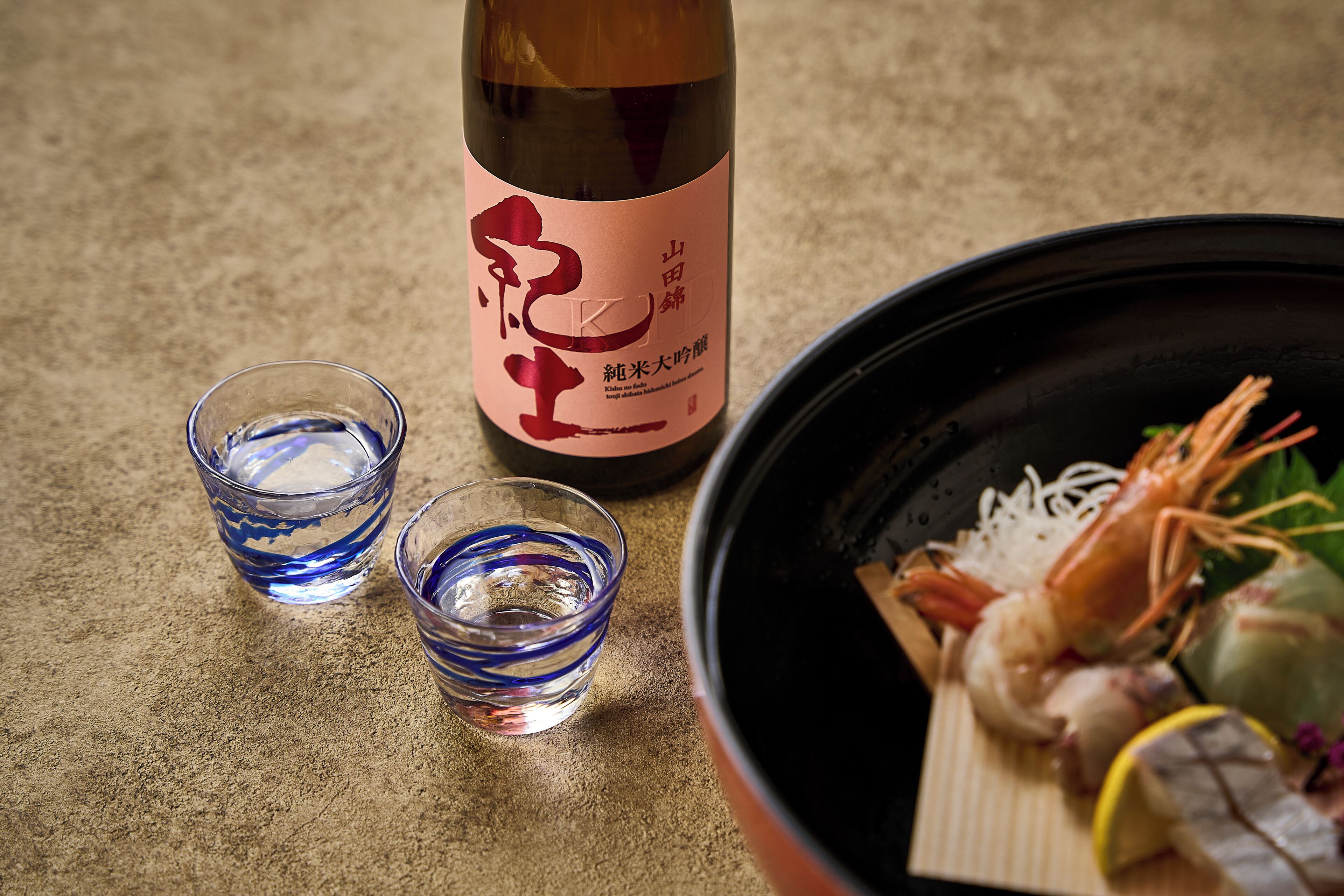 A bottle of sake, two glasses, and a bowl of seafood.