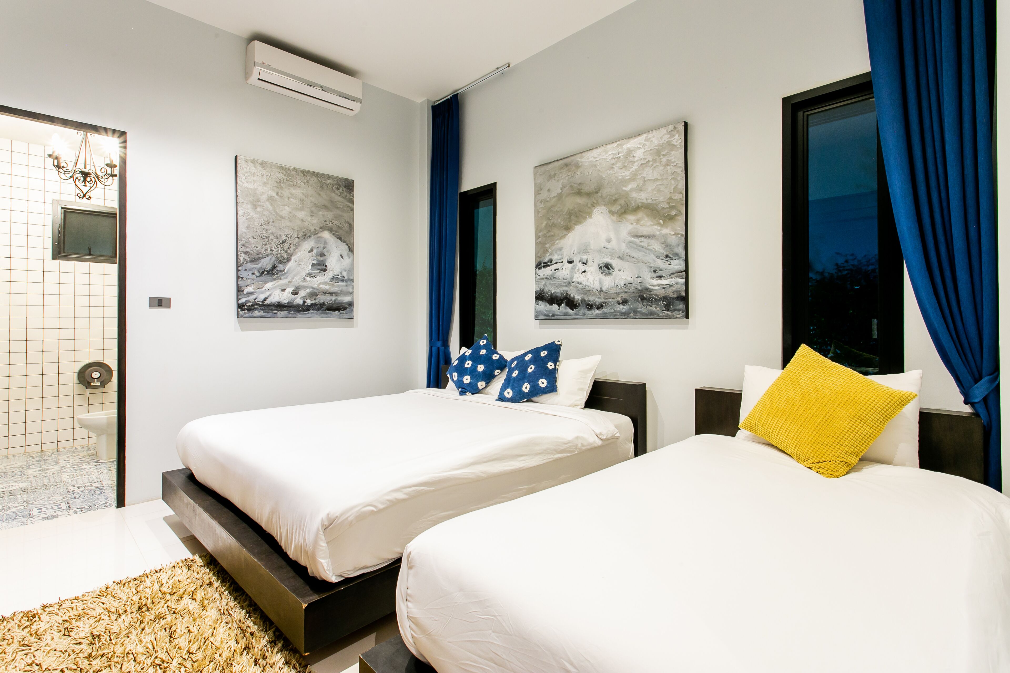 A modern hotel room with two beds, a large window with blue curtains, and artwork on the walls.