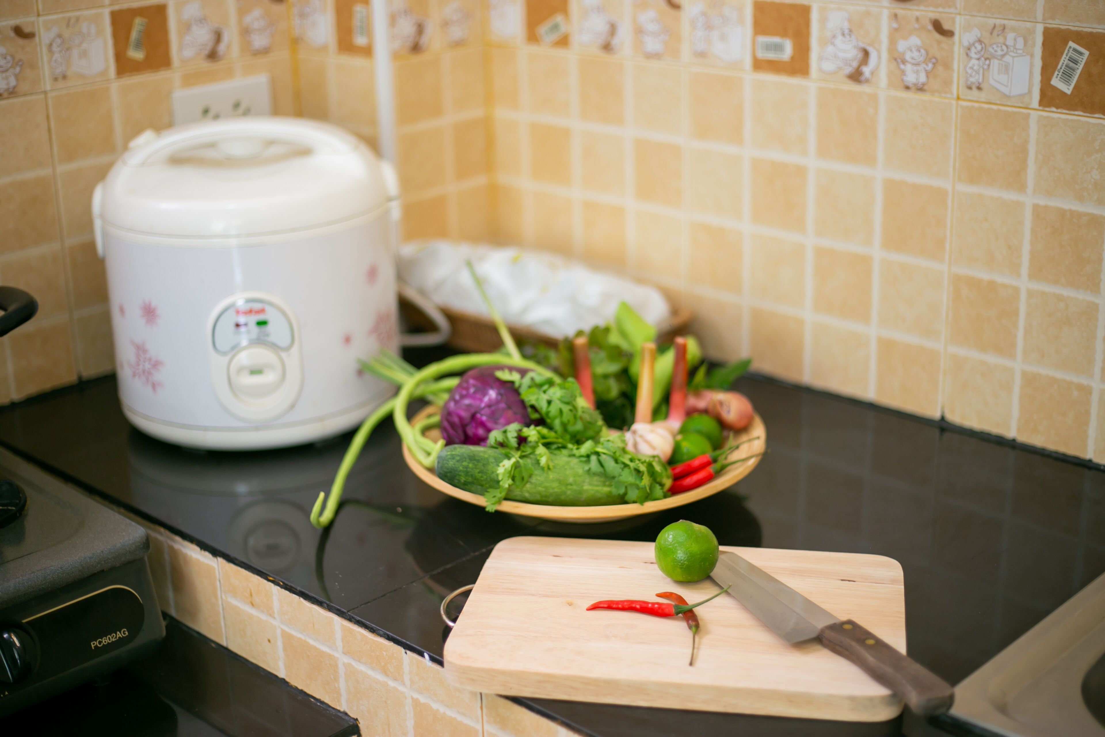 Una cocina con una cocedora de arroz, una tabla de cortar con verduras y un cuchillo.