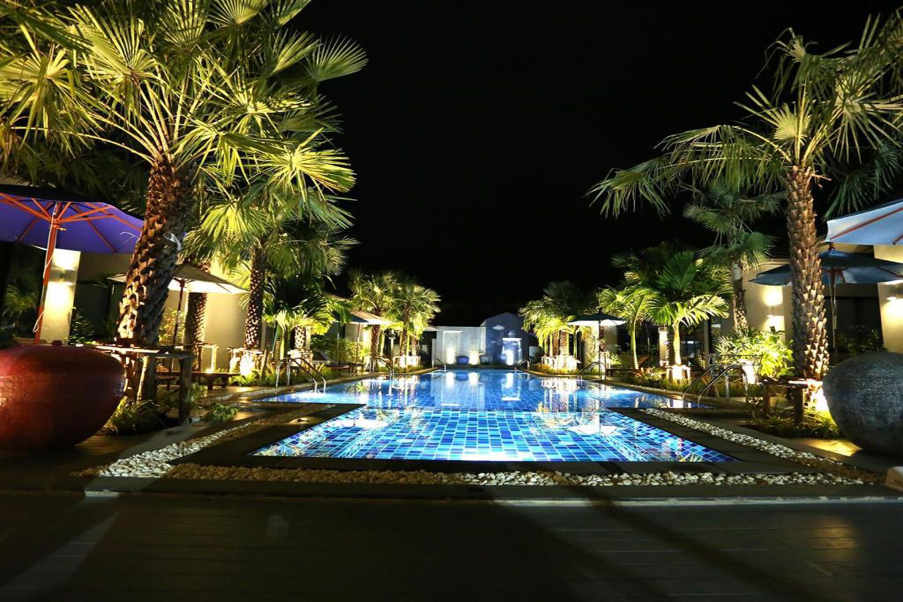 A nighttime swimming pool area with palm trees and umbrellas, surrounded by buildings.