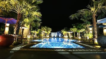 A nighttime swimming pool area with palm trees and umbrellas, surrounded by buildings.