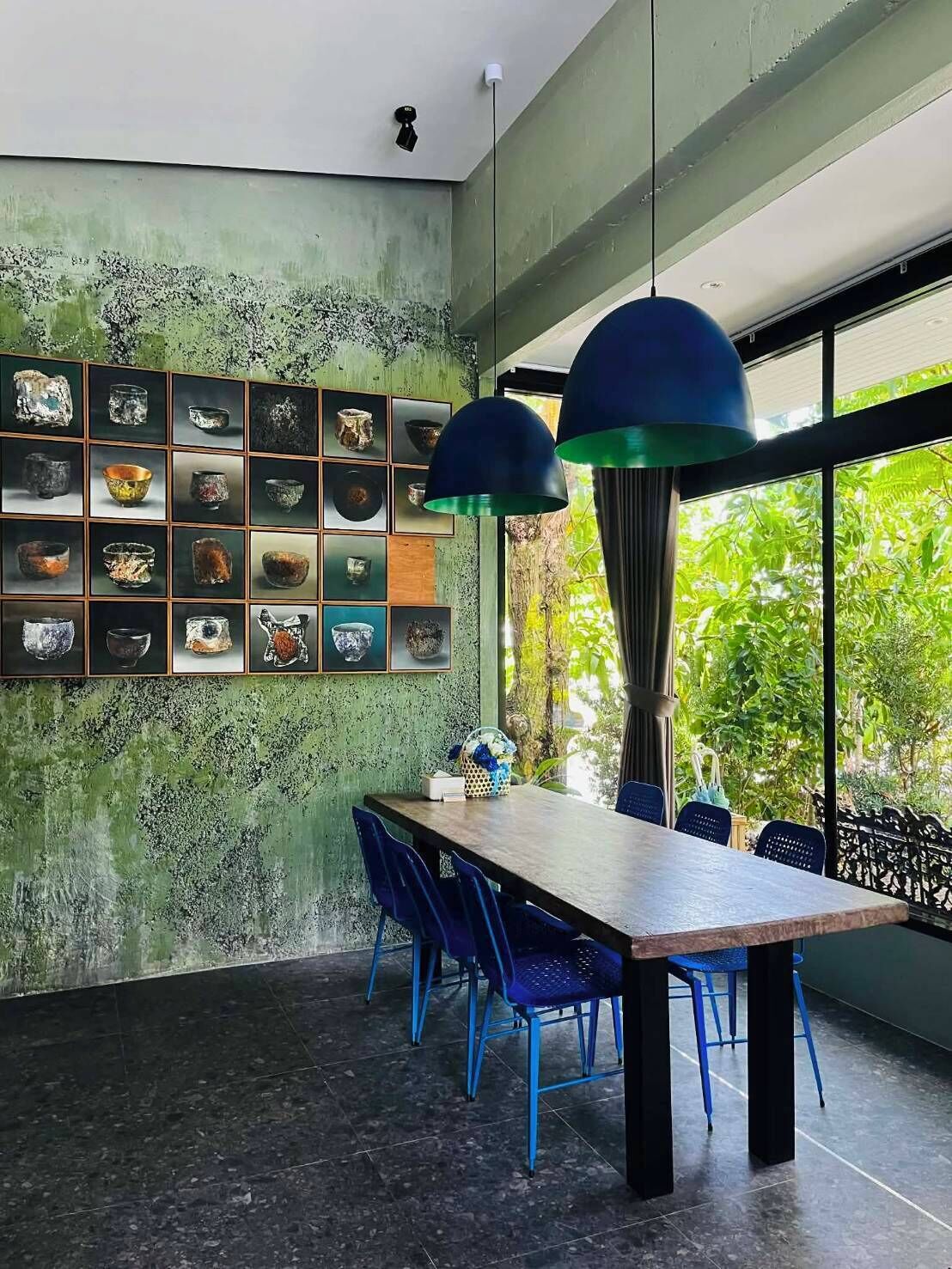 A dining area with a long table, blue chairs, a wall of framed pottery, and large windows with a view of greenery.