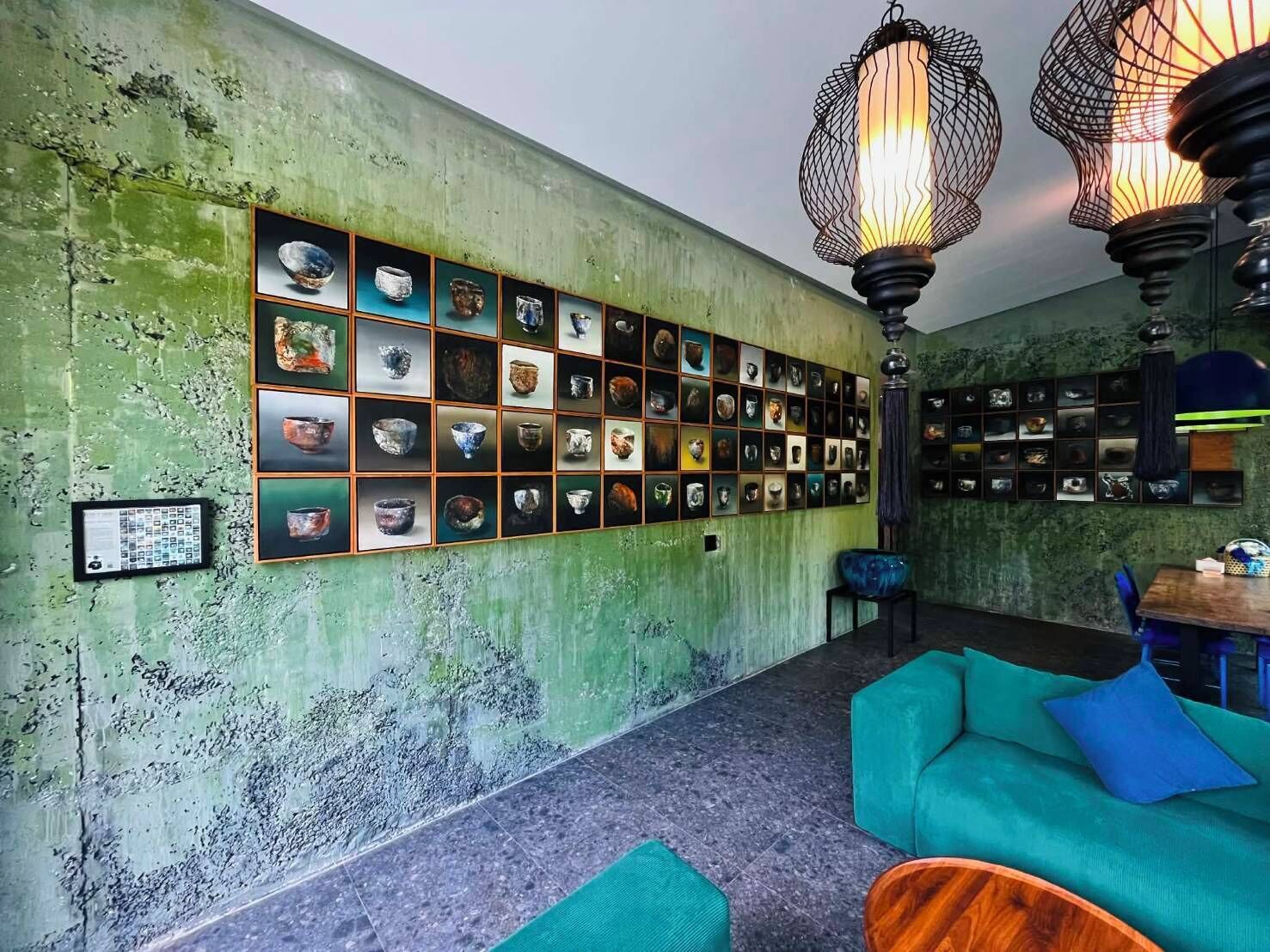 A room with a green textured wall, a wall-mounted display of various ceramic items, teal furniture, and hanging light fixtures.