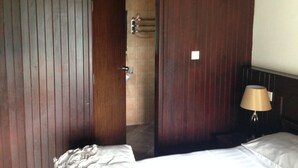 Une chambre d’hôtel avec des lambris en bois, un lit avec du linge de maison blanc, une table de chevet avec une lampe et une salle de bain visible à travers une porte ouverte.