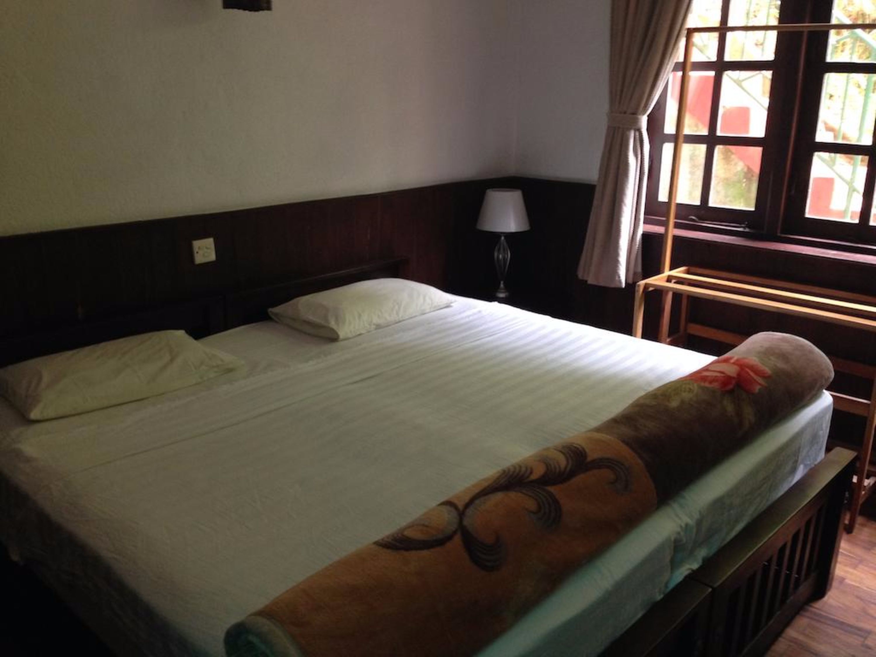 A neatly made bed with a white sheet and a decorative pillow. There is a wooden headboard, a bedside lamp, and a window with curtains.