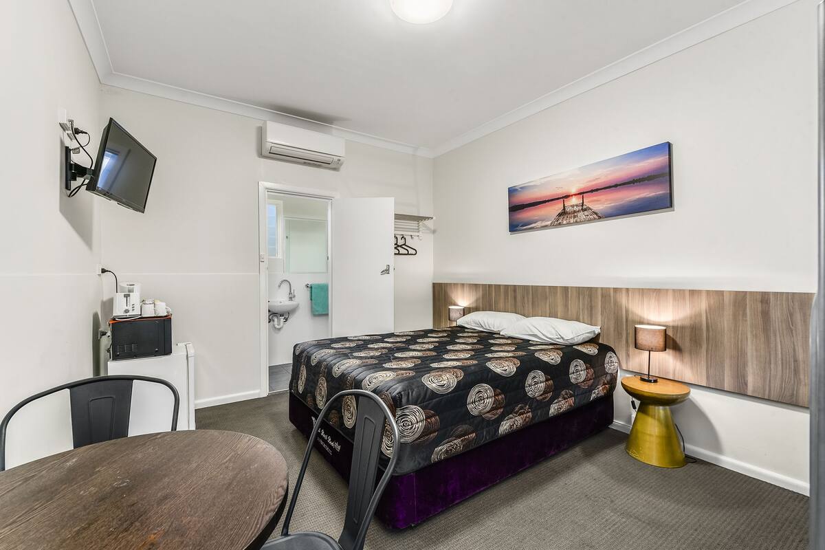 A hotel room with a bed, a desk, a chair, a TV, a painting, and a bedside lamp.