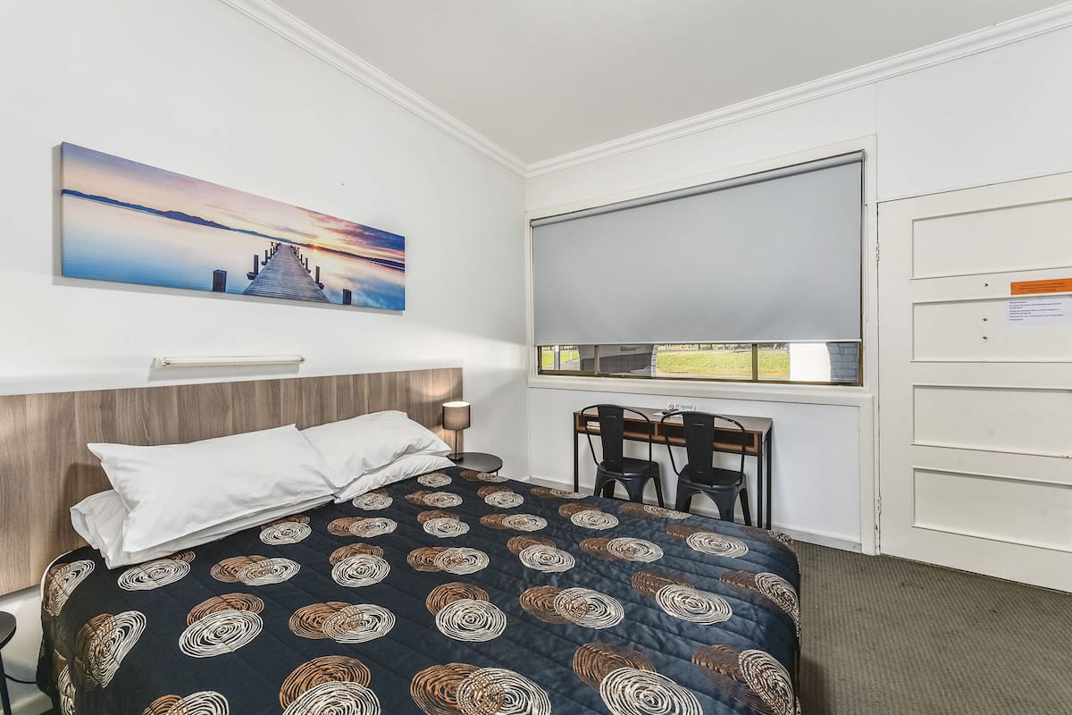 A hotel room with a bed, a desk, and a chair. There is a painting on the wall and a window with a view of the outside.