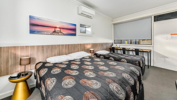 A hotel room with two beds, a desk, a chair, a lamp, an air conditioner, and a painting of a sunset.