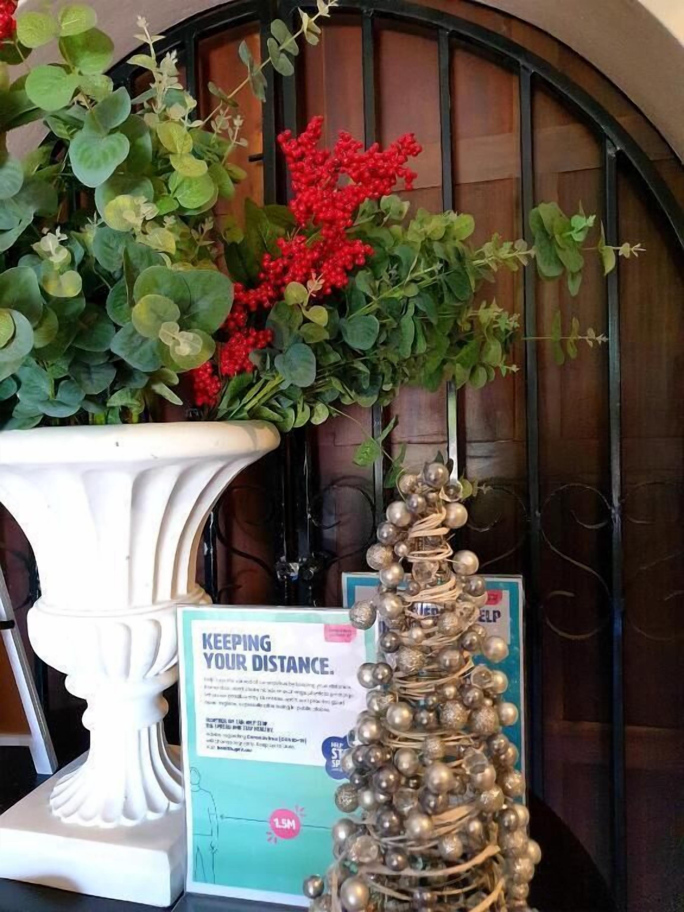A white pedestal planter with green foliage and red berries, a decorative silver ornament, and a 'KEEPING YOUR DISTANCE' sign.