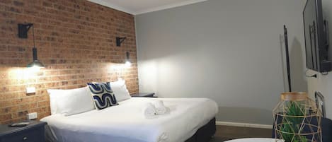 A hotel room with a brick accent wall, a bed with white bedding, a round table with a plant, and a TV mounted on the wall.